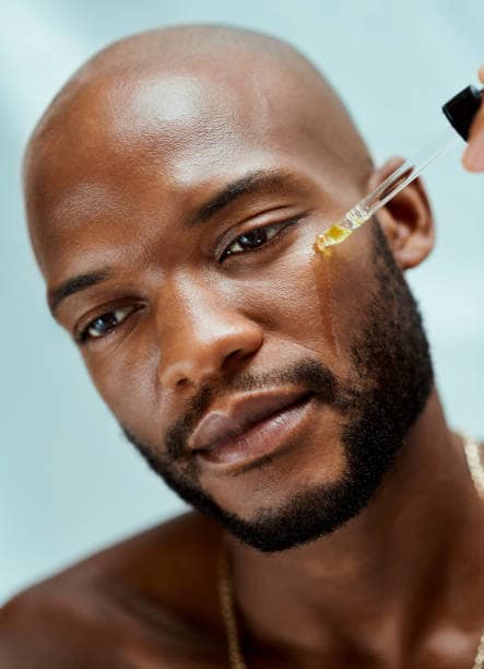 African male applying serum on his face with an oil dropper. He is doing his morning skincare routine. The beauty model has clear and clean skin